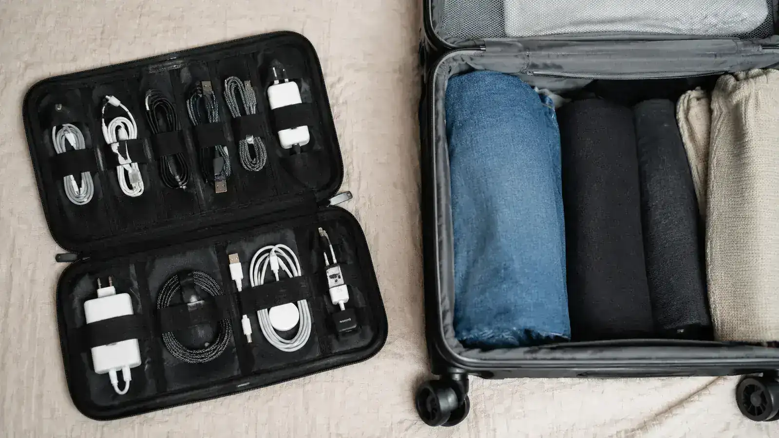 Cable organizer pouch open beside a carry-on suitcase showing all slots filled with different cables and adapters