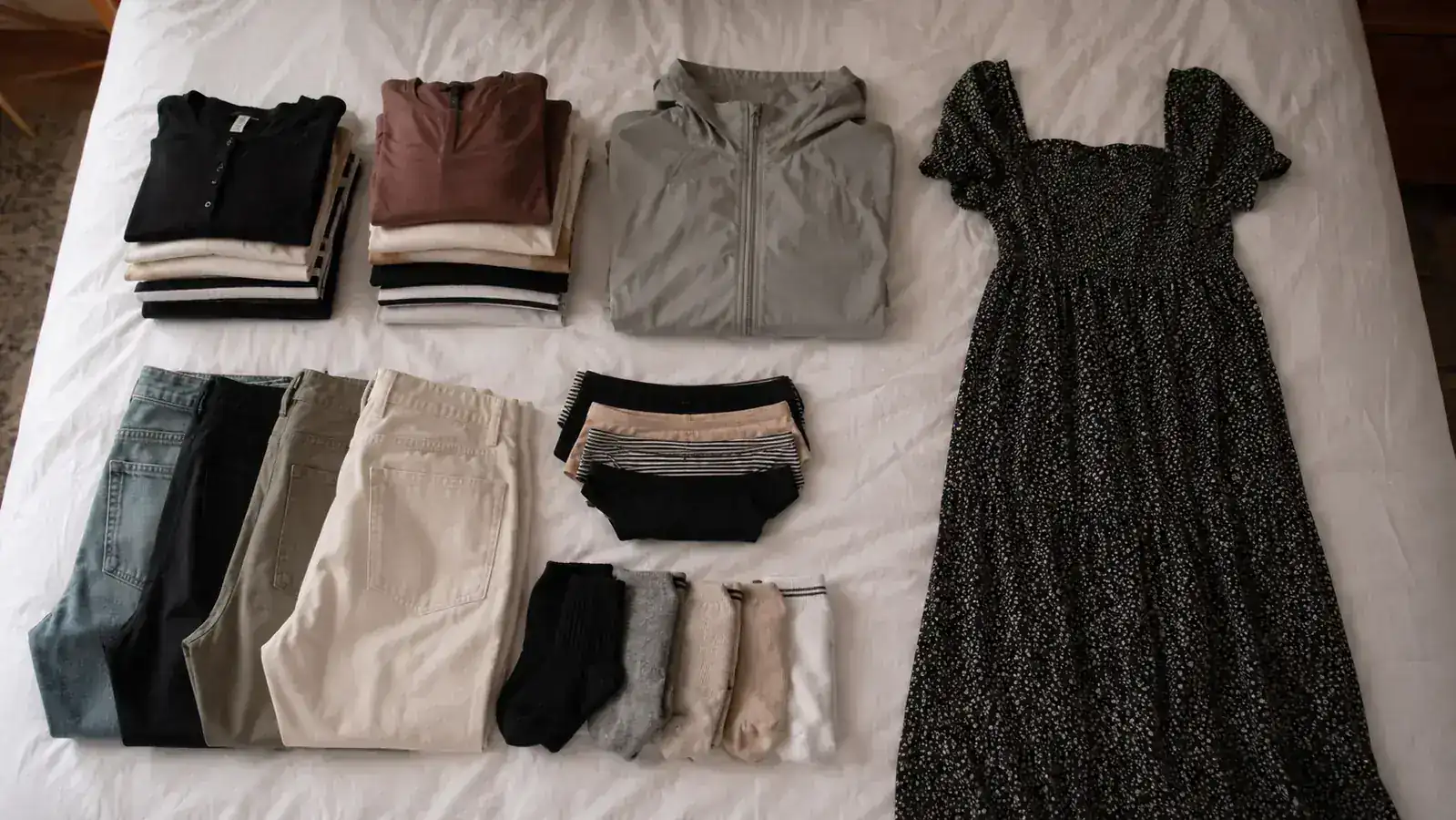 Flat lay of 14 days worth of travel clothing sorted into piles by category on a white bed